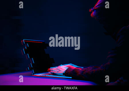Hacker in the hood working with laptop typing text in dark room. Image with glitch effect Stock Photo