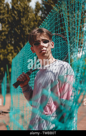 Young man outside a tennis court in Stylish urban outlook and sun glasses. The guy holds a tennis racket Stock Photo