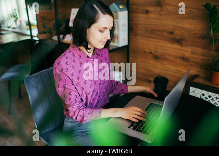 Young smiling woman working in a cafe with her laptop. One hand holding a cup with coffee, another - typing on a keyboard. Stock Photo