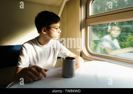 A teenager boy in glasses on a train sits at a table with tea Stock Photo