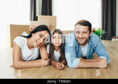 Happy family moving in new house. Mother, father and daughter together smiling lying down on floor with boxes on background Stock Photo