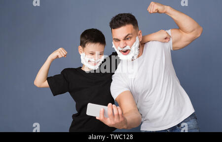 Masculine selfie. Dad and son having fun together, posing with shaving foam on cellphone camera, grey studio background Stock Photo