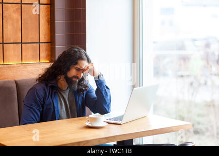 Portrait of tired thoughtful handsome young adult man freelancer in casual style sitting in cafe with laptop, trying to concentrate and remember some Stock Photo