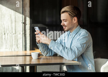 Side view portrait of handsome happy young man in blue denim shirt sitting in cafe and looking and reading message on the phone with toothy smile, loo Stock Photo