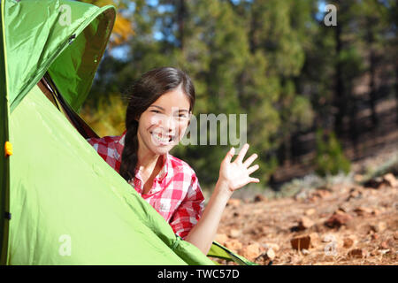 Camping woman waving hello from tent smiling happy outdoors in forest. Happy mixed race Asian Caucasian girl saying hi looking at camera. Stock Photo