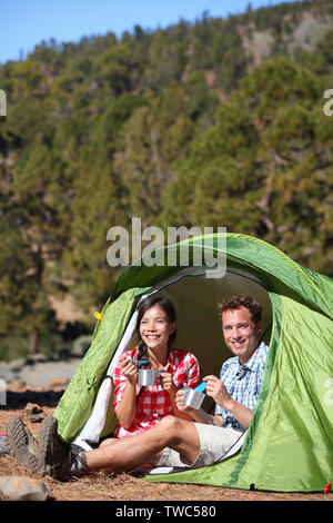 Camping people - couple eating food in tent smiling happy outdoors in forest. Happy multiracial couple having fun relaxing after outdoor activity hiking. Asian woman, Caucasian man. Stock Photo