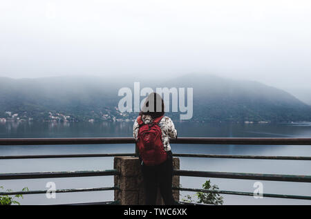 Travel girl backpacker young woman wears red Osprey rucksack at the edge of a misty blue mountain lake in Florianópolis, Brazil