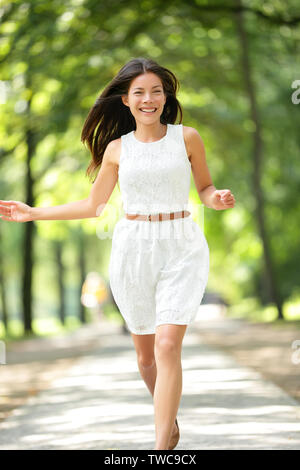 Happy Asian girl running in summer / spring park joyful and smiling in white sundress around trees. Beautiful fresh multiracial Asian Caucasian woman female model in her 20s outside. Stock Photo