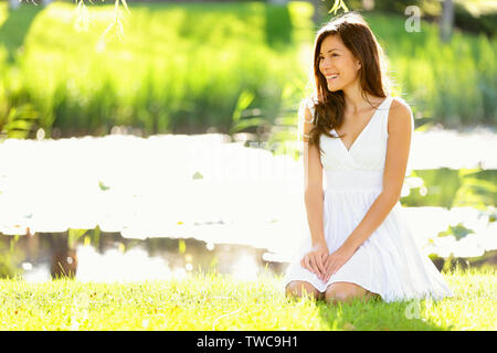 Asian woman sitting in park in spring or summer. Beautiful young woman smiling happy wearing white sundress sitting down in grass in park, Cute mixed race Asian Caucasian woman in her 20s. Stock Photo