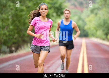 Running young mixed race couple training for marathon outside on road. Happy smiling beautiful mixed race female model in front - handsome caucasian male model blurred in background, running focused Stock Photo