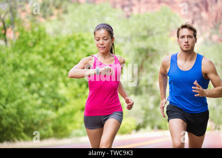 Runners - mixed multicultural couple training outside in nature. Fitness asian young smiling model checking time or pulse on heart rate monitor watch and male model jogging. Stock Photo