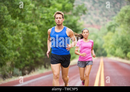 Running young fitness couple training marathon concentrated focused on road trail in summer nature. Sporty healthy mixed race woman and handsome caucasian man jogging outside. Athletic people in 20s Stock Photo