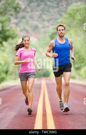 Running sporty people - two young runners jogging on road in nature training for marathon run. Multicultural couple, Asian woman sport model and man fitness model exercising together smiling happy Stock Photo