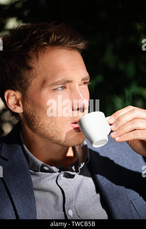 Handsome man portrait drinking espresso at cafe enjoying coffee. Close up portrait of male business man sitting outdoors drinking from espresso cup in suit jacket. Handsome modern city guy. Stock Photo