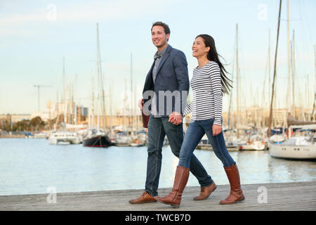 Young couple walking outdoors in old harbor, Port Vell in Barcelona Catalonia, Spain. Romantic happy woman and man holding hands enjoying life and romance outside. Multiracial Caucasian Asian couple. Stock Photo