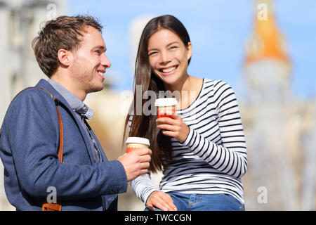 Happy couple drinking coffee talking flirting having fun together. Happy urban young man and woman laughing relaxing outside with disposable coffee cup. Caucasian Asian Interracial couple. Stock Photo