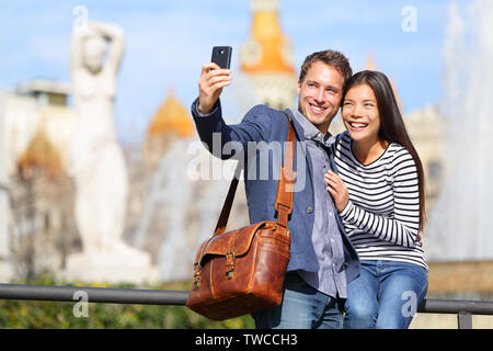 Happy urban city couple on travel in Barcelona taking selfie self portrait photograph with smart phone camera. Happy young man and woman on Placa de Catalunya, Catalonia Square, Barcelona, Spain. Stock Photo
