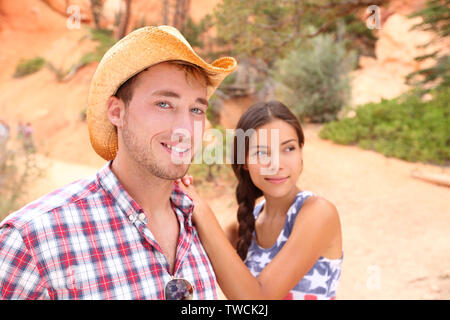 Couple portrait in american countryside outdoors. Smiling multiracial young couple in western USA nature. Man wearing cowboy hat and woman wearing USA flag shirt. Stock Photo