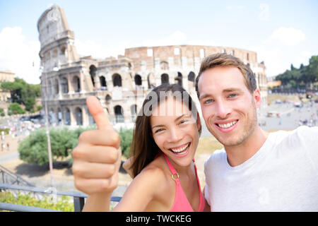 Selfie - Romantic travel couple by Coliseum, Rome, Italy. Happy lovers on honeymoon sightseeing having fun in front of Colosseum. Woman giving thumbs up in tourism travel concept. Stock Photo