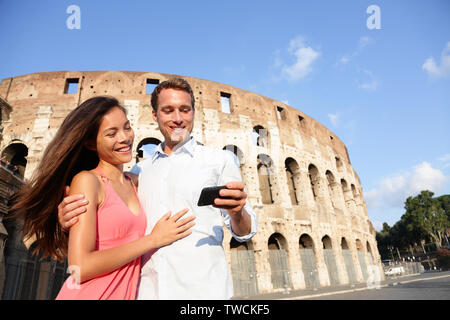 Couple in Rome by Colosseum using smart phone looking at pictures or using travel app in Italy. Happy lovers on honeymoon sightseeing Coliseum. Love and travel concept with multiracial couple. Stock Photo