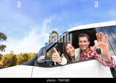 New car - happy couple showing car keys driving having fun on road trip drive in rental car. Happy lifestyle with beautiful young interracial couple outdoors on travel. Man driver and woman passenger. Stock Photo