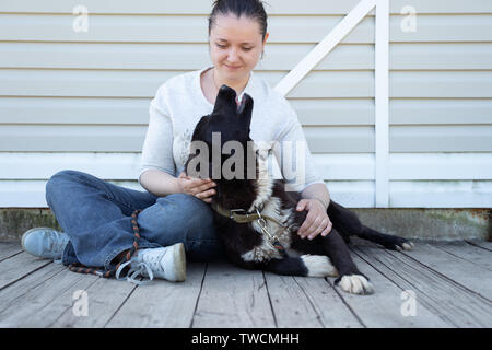 Picture of seated brunette woman in jeans and white jacket stroking black dog on white background wooden wall Stock Photo