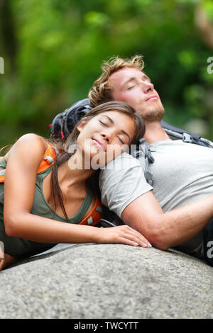 Hiking couple lovers relaxing sleeping in nature. Tried hikers resting lying down outdoors taking a break from hike. Young Asian woman and Caucasian man. Stock Photo