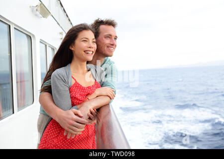 Cruise ship couple romantic enjoying travel on boat embracing looking at view. Happy lovers traveling on vacation sailing on open sea ocean enjoying romance. Young Asian woman and Caucasian man. Stock Photo