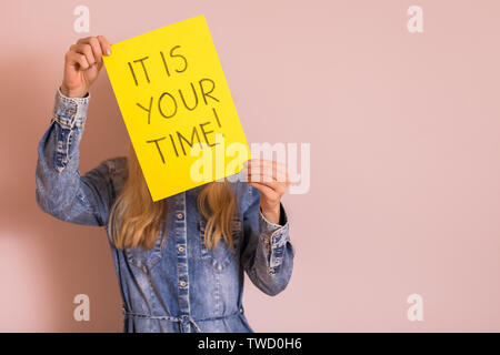 Woman holding paper with text it is your time while standing in front of the wall. Stock Photo