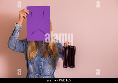 Woman holding empty wallet and sad face on paper while standing in front of wall. Stock Photo