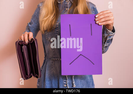 Woman holding empty wallet and sad face on paper while standing in front of wall. Stock Photo