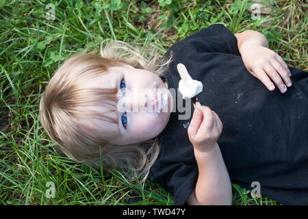 top view of little girl with chocolate popsicle ice cream in the hand lying on green grass closeup Stock Photo