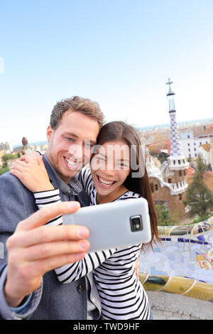 Happy travel couple talking selfie self-portrait with smartphone in Park Guell, Barcelona, Spain. Beautiful young multiracial couple looking at camera taking photo with smart phone smiling in love. Stock Photo