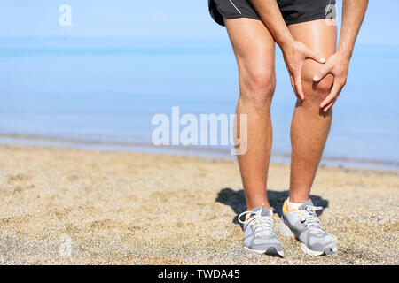 Running injury - Man jogging with knee pain. Close-up view of runner injured jogging on the beach clutching his knee in pain. Male fitness athlete. Stock Photo