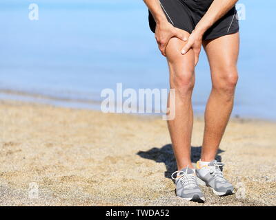 Muscle injury. Runner man with sprain thigh muscle. Athlete in sports shorts clutching his thigh muscles after pulling or straining them while jogging on the beach wearing running shoes. Stock Photo