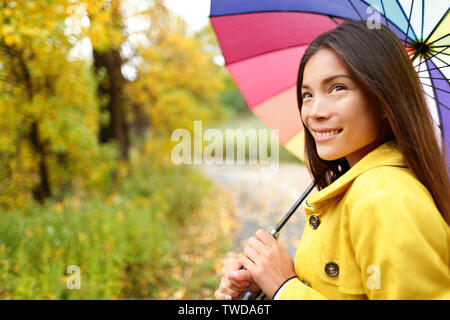 Woman happy with umbrella under the rain during Autumn forest walk. Girl enjoying rainy fall day looking away smiling. Mixed race Caucasian / Asian chinese girl. Stock Photo