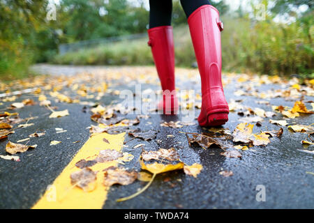 Autumn fall concept with colorful leaves and rain boots outside. Close up of woman feet walking in red boots. Stock Photo