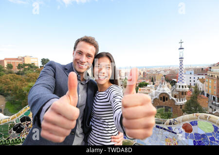 Travel couple happy in Park Guell, Barcelona, Spain. Beautiful young multiracial couple giving thumbs up hand sign smiling happy having fun on Europe vacation trip. Asian woman, Caucasian man. Stock Photo