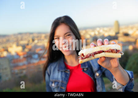 Jamon Iberico Sandwich. Woman eating in Barcelona, Spain showing traditional Spanish food, cured Serrano ham. Focus on bocadillo, Barcelona skyline in background. Stock Photo