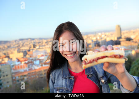 Jamon Serrano Sandwich. Woman eating in Barcelona, Spain showing traditional Spanish food, cured Iberico ham. Focus on girl, Barcelona skyline in background. Stock Photo