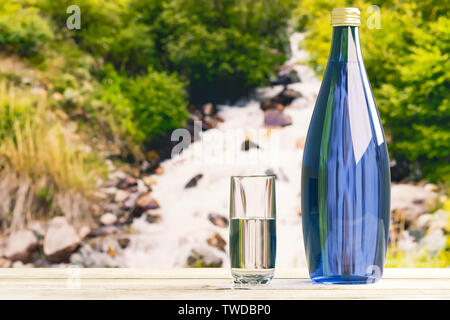 A bottle and a glass with fresh cool water against the backdrop of nature, a mountain river with the purest water. Stock Photo