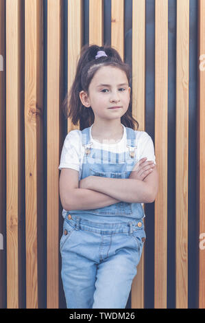 Cute happy caucasian girl in jeans overalls on a background of wall with wooden vertical slats. Lifestyle Stock Photo