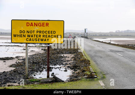 The Danger warning sign for tourist traffic crossing the 3-mile stretch of tarmac road at low tide connecting Lindisfarne Holy Island from the mainland Stock Photo