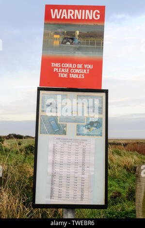 A warning sign and a tide table guide on the causeway to Lindisfarne Holy Island in Northumberland, Britain. The small tidal island is only accessible Stock Photo