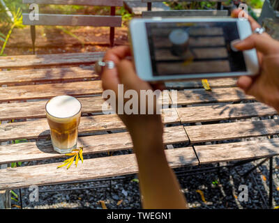 Woman's hands holding white smartphone and taking a photo of iced cappuccino coffee in glass on wooden table top view Stock Photo