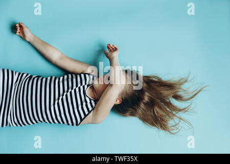 Girl laying on blue background covering face. Child sleeping flat lay top view. Having rest, relaxing. Childhood memories. Beautiful flowing hair. Simple minimalist background, wallpaper Stock Photo