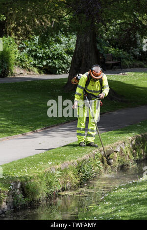 A Cormac worker strimming grass and weeds around a stream in a landscaped park in Newquay in Cornwall.