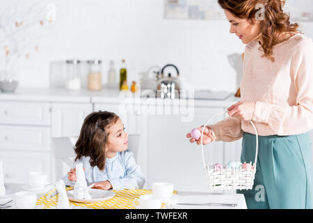 Curly woman holding wicker basket with easter eggs and looking at daughter in kitchen Stock Photo