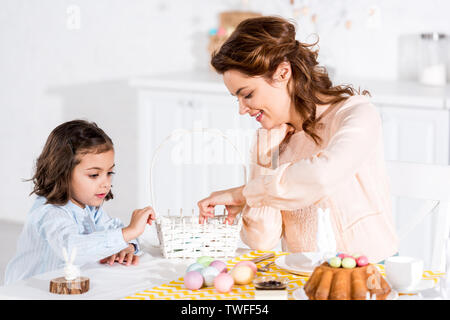 Mother and daughter putting easter eggs in wicker basket in kitchen Stock Photo