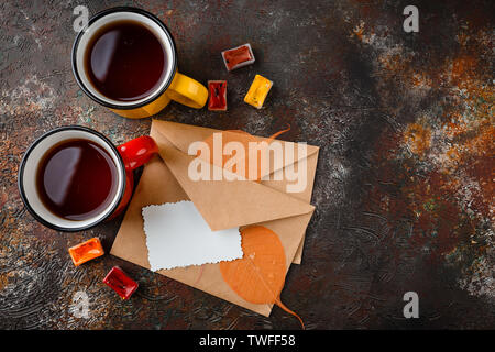 Two colored enamel cups of tea, two envelops, watercolors in cuvettes, , autumn leaves and bumps on a rusty brown background Stock Photo